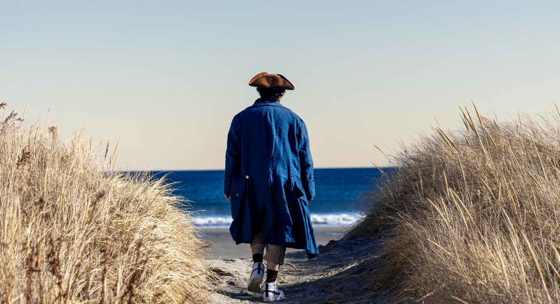 Person in colonial-style clothing walking toward the ocean between sand dunes.